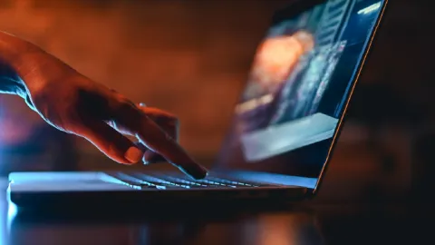 Close-up of a person’s hand typing on a laptop keyboard in low light with a warm orange glow in the background.